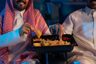 شاشات ضوئية كبيرة لزيادة الاستمتاع بالتجربة، القيام بنشاطات ترفيهية ممتعة, الاستمتاع بتناول المقرمشات ذهبية اللون ، مشاهدة الأفلام والمسرحيات والعروض المتنوعة, رجلان عربيان خليجيان سعوديان يجلسان في قاعة السينما ويتابعان الفيلم بايماءات السعادة, دور العرض الحديثة في المملكة العربية السعودية