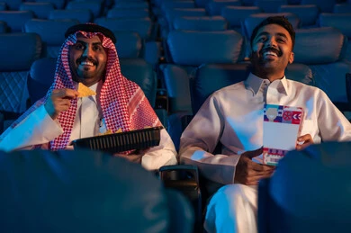 دور العرض الحديثة في المملكة العربية السعودية، القيام بنشاطات ترفيهية ممتعة, رجلان عربيان خليجيان سعوديان يجلسان في قاعة السينما ويتابعان  الفيلم بايماءات السعادة, مشاهدة الأفلام والمسرحيات والعروض المتنوعة, شاشات ضوئية كبيرة لزيادة الاستمتاع بالتجربة