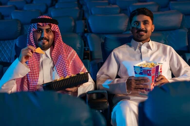 رجلان عربيان خليجيان سعوديان يجلسان في قاعة السينما ويتابعان  الفيلم بايماءات السعادة,القيام بنشاطات ترفيهية ممتعة, مشاهدة الأفلام والمسرحيات والعروض المتنوعة, شاشات ضوئية كبيرة لزيادة الاستمتاع بالتجربة، دور العرض الحديثة في المملكة العربية السعودية