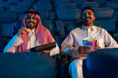 شاشات ضوئية كبيرة لزيادة الاستمتاع بالتجربة، القيام بنشاطات ترفيهية ممتعة, رجلان عربيان خليجيان سعوديان يجلسان في قاعة السينما ويتابعان  الفيلم بايماءات السعادة, مشاهدة الأفلام والمسرحيات والعروض المتنوعة, دور العرض الحديثة في المملكة العربية السعودية