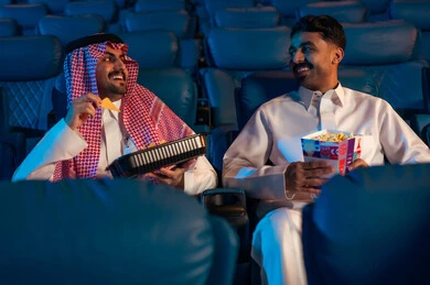 Engaging in enjoyable recreational activities, two Saudi Arabian Gulf men are sitting in a cinema hall watching the movie with gestures of happiness, watching films, plays, and various shows, large screens to enhance the enjoyment of the experience, modern theaters in the Kingdom of Saudi Arabia.