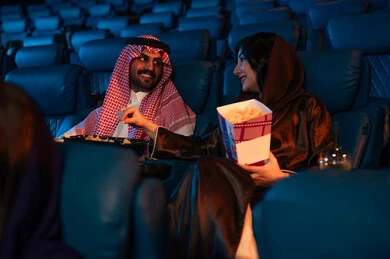 4D cinema halls, happy recreational activities, spending enjoyable times, a Saudi Arabian Gulf man next to his wife in the cinema hall, expressions indicating engagement, enjoying popcorn and cold drinks, watching movies and theatrical performances in the cinema, cinema and theater, background of the cinema hall.