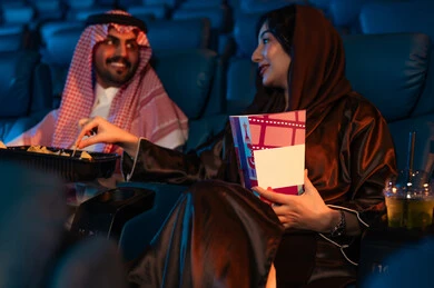 The concept of affection and companionship, happy recreational activities, spending enjoyable times, an Arab Saudi Gulf man next to his wife in a cinema hall, expressions indicating integration, enjoying popcorn and cold drinks, watching movies and theatrical performances in the cinema, cinema and theater, background of the cinema screening room, exhibition hall.