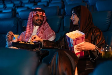 Enjoying popcorn, happy recreational activities, spending enjoyable times, an Arab Saudi Gulf man next to his wife in the cinema hall, expressions indicating engagement, watching movies and theatrical performances in the cinema, cinema and theater, background of the cinema screening room, exhibition hall.