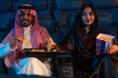 Engaging in fun recreational activities, spending happy times, an Arab Saudi Gulf man next to his wife in the cinema hall, expressions indicating immersion, enjoying popcorn and cold drinks, watching movies and theatrical performances in the cinema, cinema and theater, background of the cinema screening room, exhibition hall.