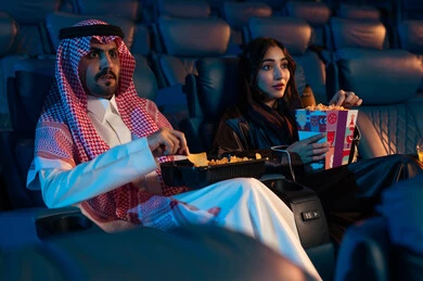 A Saudi Arabian Gulf man next to his wife in a cinema hall, happy recreational activities, spending enjoyable times, expressions indicating engagement, enjoying popcorn and cold drinks, watching movies and theatrical performances in the cinema, cinema and theater, background of the cinema screening room, exhibition hall.