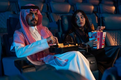 Expressions of joy and happiness, happy recreational activities, spending enjoyable times, an Arab Saudi Gulf man next to his wife in the cinema hall, expressions indicating engagement, enjoying popcorn and cold drinks, watching movies and theatrical performances in the cinema, cinema and theater, background of the cinema screening room, exhibition hall.