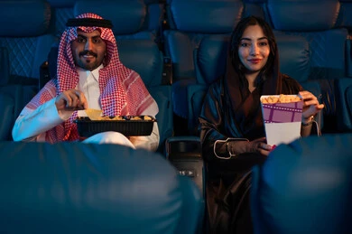 Spending enjoyable times, happy recreational activities, an Arab Saudi Gulf man next to his wife in the cinema hall, expressions indicating engagement, enjoying popcorn and cold drinks, watching movies and theatrical performances in the cinema, cinema and theater, background of the cinema screening room, exhibition hall.