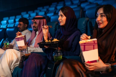 Engaging in fun recreational activities, an Arab Saudi Gulf audience in the cinema hall, expressions indicating immersion, enjoying popcorn and cold drinks, spending enjoyable times, watching movies and theatrical performances in the cinema, cinema and theater, background of the cinema screening room, screening hall.