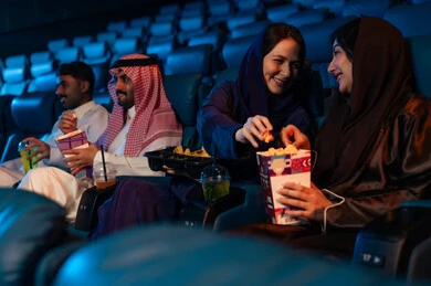 Spending enjoyable times, an Arab Saudi Gulf audience in the cinema hall, expressions indicating engagement, enjoying popcorn and cold drinks, recreational activities, watching movies and theatrical performances in the cinema, cinema and theater, background of the cinema screening room, the screening hall.