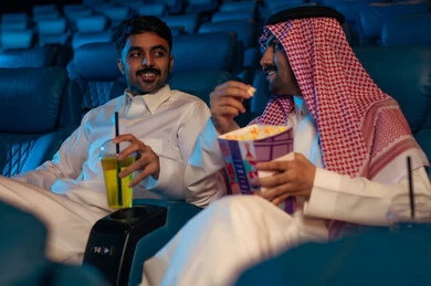 Engaging in enjoyable recreational activities, two Saudi Arabian Gulf men are sitting in a cinema hall watching the movie with gestures of happiness, watching films, plays, and various shows, large screens to enhance the enjoyment of the experience, modern theaters in the Kingdom of Saudi Arabia.