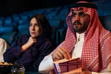 Expressions of fear and terror, excitement and suspense and immersion, spending enjoyable times, an Arab Saudi Gulf man in the cinema hall with his wife, enjoying popcorn and cold drinks, watching horror movies and theatrical performances in the cinema, cinema and theater, background of the cinema screening room, exhibition hall.