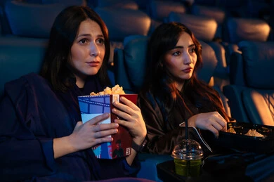Enjoying popcorn and snacks, watching horror movies and theatrical performances in the cinema, expressions of fear and terror, excitement and suspense and immersion, spending enjoyable times, two Arab Saudi Gulf friends in the cinema hall, cinema and theater, background of the cinema screening room, exhibition hall.