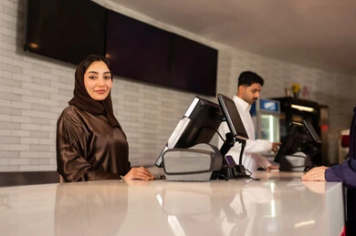 A smiling Saudi Arabian Gulf receptionist wearing an abaya and hijab is looking at the camera in the ticket booking area to watch a movie in the cinema hall, watching films and theatrical performances, and enjoying distinguished recreational activities.