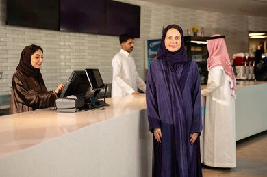 مشاهدة الأفلام والعروض المسرحية ، امراه عربية سعودية خليجية مبتسمه ترتدي العباءه والحجاب تنظر الى الكاميرا فى صاله حجز التذاكر لمشاهدة فيلم في صالة السينما،  أنشطه ترفيهيه متميزة