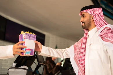 أنشطه ترفيهيه متميزة، رجل عربي سعودي خليجي مبتسم يرتدي الزي التقليدي والشماغ يقوم بشراء الفشار فى صاله حجز التذاكر لمشاهدة فيلم في صالة السينما، متابعة الأفلام والعروض المسرحية