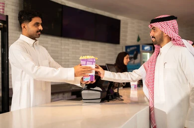 متابعة الأفلام والعروض المسرحية ، رجل عربي سعودي خليجي مبتسم يرتدي الزي التقليدي والشماغ يقوم بشراء الفشار فى صاله حجز التذاكر لمشاهدة فيلم في صالة السينما، أنشطه ترفيهيه متميزة