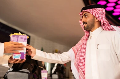 رجل عربي سعودي خليجي مبتسم يرتدي الزي التقليدي والشماغ يقوم بشراء الفشار فى صاله حجز التذاكر لمشاهدة فيلم في صالة السينما، متابعة الأفلام والعروض المسرحية ، أنشطه ترفيهيه متميزة