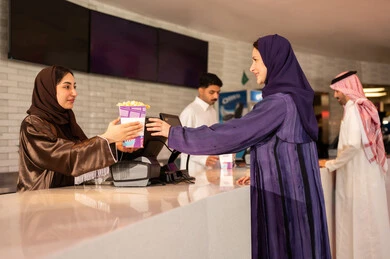 سيدة عربية سعودية خليجية مبتسمة ترتدي العباءة والحجاب تقوم بشراء الفشار فى صاله حجز التذاكر لمشاهدة فيلم في صالة السينما، متابعة الأفلام والعروض المسرحية ، أنشطه ترفيهيه متميزة