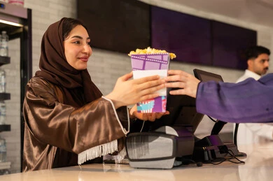 الاستمتاع بتناول الفشار ،سيدة عربية سعودية خليجية مبتسمة ترتدي العباءة والحجاب تقوم بشراء الفشار فى صاله حجز التذاكر لمشاهدة فيلم في صالة السينما، متابعة الأفلام والعروض المسرحية ، أنشطه ترفيهيه متميزة