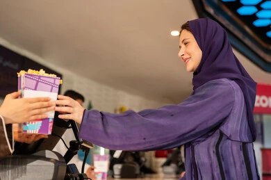 متابعة الأفلام والعروض المسرحية ،سيدة عربية سعودية خليجية مبتسمة ترتدي العباءة والحجاب تقوم بشراء الفشار فى صاله حجز التذاكر لمشاهدة فيلم في صالة السينما،  أنشطه ترفيهيه متميزة