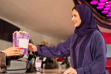 تجربة سينمائية خيالية ، سيدة عربية سعودية خليجية مبتسمة ترتدي العباءة والحجاب تقوم بشراء الفشار فى صاله حجز التذاكر لمشاهدة فيلم في صالة السينما، متابعة الأفلام والعروض المسرحية ، أنشطه ترفيهيه متميزة