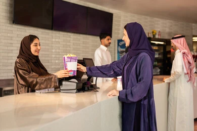 سيدة عربية سعودية خليجية مبتسمة ترتدي العباءة والحجاب تقوم بشراء الفشار فى صاله حجز التذاكر لمشاهدة فيلم في صالة السينما، متابعة الأفلام والعروض المسرحية ، أنشطه ترفيهيه متميزة