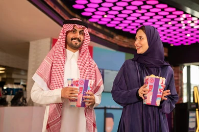 رجل عربي خليجي سعودي يرتدي الزى التقليدي والشماغ برفقة زوجته يمسكان بيدهم كيس الفشار فى صاله حجز التذاكر لمشاهدة فيلم في صالة السينما، متابعة الأفلام والعروض المسرحية ، أنشطه ترفيهيه متميزة
