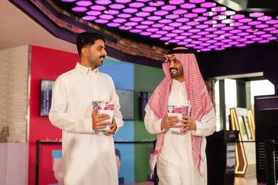 Watching movies and theatrical performances, two smiling Arab Gulf men from Saudi Arabia wearing traditional attire and shemagh are holding a bag of popcorn in the ticket booking area to watch a movie in the cinema hall, distinctive recreational activities.