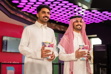 Distinctive recreational activities, two smiling Arab Gulf men from Saudi Arabia wearing traditional attire and shemagh are holding a bag of popcorn in the ticket booking area to watch a movie in the cinema hall, following films and theatrical performances. Distinctive recreational activities, two smiling Arab Gulf men from Saudi Arabia wearing traditional attire and shemagh are holding a bag of popcorn in the ticket booking area to watch a movie in the cinema hall, following films and theatrical performances.