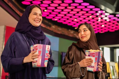 صديقتان عربيتان خليجيتان سعوديتان مبتسمتان ترتديان العباءه والحجاب  تمسكان بيدهم كيس الفشار فى صاله حجز التذاكر لمشاهدة فيلم في صالة السينما، متابعة الأفلام والعروض المسرحية ، أنشطه ترفيهيه متميزة