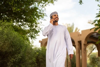 الرد على الاتصالات والمكالمات، استخدام الأجهزة الحديثة والمتطورة, رجل عربي خليجي عماني يرتدي الدشداشة والكمه يقوم بالمشي فى احد الحدائق يمسك بيده الجوال، التواصل مع الأهل والأصدقاء عبر الهاتف المحمول، قضاء وقت ممتع فى الخارج