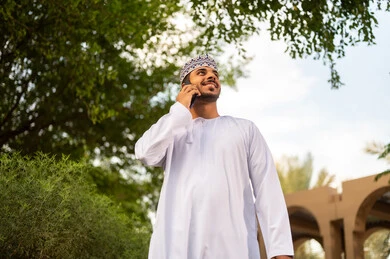 استخدام الأجهزة الحديثة والمتطورة، الرد على الاتصالات والمكالمات, رجل عربي خليجي عماني يرتدي الدشداشة والكمه يقوم بالمشي فى احد الحدائق يمسك بيده الجوال، التواصل مع الأهل والأصدقاء عبر الهاتف المحمول، قضاء وقت ممتع فى الخارج