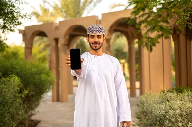 رجل عربي خليجي عماني يرتدي الدشداشة و الكمه يمسك بيده هاتف محمول بشاشة سوداء فارغة، عرض خدمات وتطبيقات الهاتف المحمول، التسوق الإلكتروني، ايماءات وجه سعيدة، قضاء عكله نهايه الاسبوع فى احدي الحدائق ، الاستمتاع بالجو الهاديء