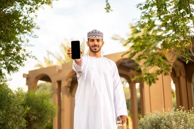 An Arab Gulf Omani man wearing a dishdasha and a kumma is holding a mobile phone with a blank black screen, showcasing mobile services and applications, online shopping, happy facial expressions, spending the weekend in one of the parks, enjoying the calm atmosphere.