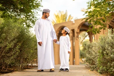 Family lifestyle, spending enjoyable times with children in parks and public gardens, an Omani Gulf Arab boy wearing a dishdasha and a kumma holding his father's hand while strolling in the park, spending the weekend.
