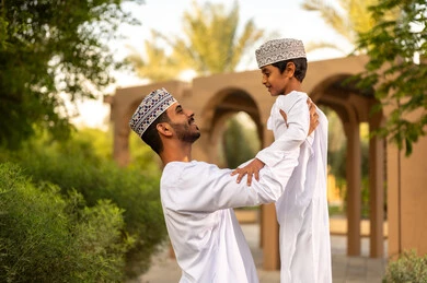 الإستمتاع بالفعاليات الصيفية، قضاء أوقات عائلية ممتعة ومسلية خلال العطلة, أب عربي خليجي عماني يرتدي الدشداشة و الكمة يقوم باللعب مع ابنه ويرفعه عاليا في الهواء بايماءات السعادة و السرور