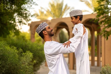 قضاء أوقات عائلية ممتعة ومسلية خلال العطلة, أب عربي خليجي عماني يرتدي الدشداشة و الكمة يقوم باللعب مع ابنه ويرفعه عاليا في الهواء بايماءات السعادة و السرور , الإستمتاع بالفعاليات الصيفية