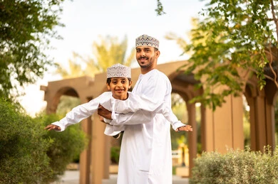 الأنشطة المرحة للأبناء والعائلة في وقت الفراغ, أب عربي خليجي عماني يرتدي الدشداشة و الكمة يقوم باللعب مع ابنه ويرفعه عاليا في الهواء بايماءات السعادة و السرور , الإستمتاع بالفعاليات الصيفية