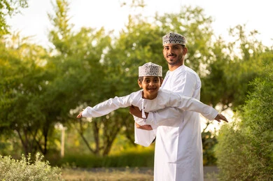 الترفيه والمرح العائلي في وقت الإجازة, أب عربي خليجي عماني يرتدي الدشداشة و الكمة يقوم باللعب مع ابنه ويرفعه عاليا في الهواء بايماءات السعادة و السرور , الإستمتاع بالفعاليات الصيفية