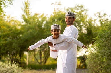 المرح العائلي أثناء الإجازة, أب عربي خليجي عماني يرتدي الدشداشة و الكمة يقوم باللعب مع ابنه ويرفعه عاليا في الهواء بايماءات السعادة و السرور , الإستمتاع بالفعاليات الصيفية