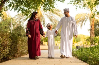المرح والتسلية في الجو العائلي, أنشطة ترفيهية عائلية في العطلة، عائله عربية خليجيه عمانية ترتدي الزي التقليدي تقوم بالمشي فى احدي الحدائق برفقة ابنهم بايماءات السعادة و السرور