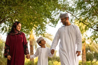 أوقات سعيدة مع أفراد الأسرة, أنشطة ترفيهية عائلية في العطلة، عائله عربية خليجيه عمانية ترتدي الزي التقليدي تقوم بالمشي فى احدي الحدائق برفقة ابنهم بايماءات السعادة و السرور