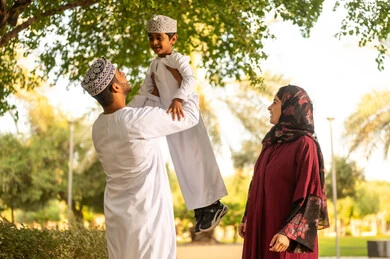 الاستمتاع بالنشاطات الخارجية، اب عربي خليجي عماني يرتدي الدشداشة و الكمة يقوم برفع ابنه الي الاعلى برفقه زوجته بايماءات السعادة و السرور ، قضاء وقت ممتع مع الابناء