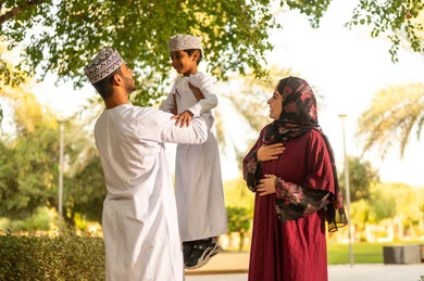 الخروج للتمتع بالنزهة والمساحات الخضراء، اب عربي خليجي عماني يرتدي الدشداشة و الكمة يقوم برفع ابنه الي الاعلى برفقه زوجته بايماءات السعادة و السرور ، قضاء وقت ممتع مع الابناء
