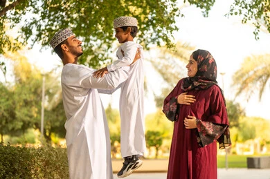 قضاء لحظات مرحة وسط الطبيعة، اب عربي خليجي عماني يرتدي الدشداشة و الكمة يقوم برفع ابنه الي الاعلى برفقه زوجته بايماءات السعادة و السرور ، قضاء وقت ممتع مع الابناء فى الخارج