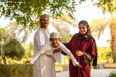 التنزه والتمتع بالهواء النقي، اب عربي خليجي عماني يرتدي الدشداشة و الكمة يقوم برفع ابنه الي الاعلى برفقه زوجته بايماءات السعادة و السرور ، قضاء وقت ممتع مع الابناء فى الخارج