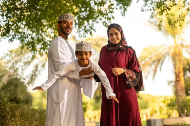 الترفيه والمرح خارج المنزل، اب عربي خليجي عماني يرتدي الدشداشة و الكمة يقوم برفع ابنه الي الاعلى برفقه زوجته بايماءات السعادة و السرور ، قضاء وقت ممتع مع الابناء فى الخارج
