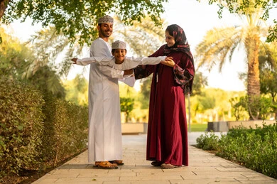 الاستمتاع بالتمشية في الهواء الطلق، اب عربي خليجي عماني يرتدي الدشداشة و الكمة يقوم برفع ابنه الي الاعلى برفقه زوجته بايماءات السعادة و السرور ، قضاء وقت ممتع مع الابناء فى الخارج