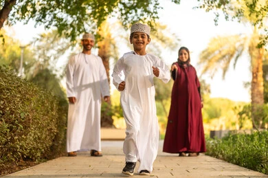 الإستمتاع باللعب في يوم مشمس، عائلة عربية خليجية عمانية يرتدون الزي التقليدي يمشون في إحدى الحدائق العامة في النهار، تقوية وتعزيز الروابط الأسرية، الاستمتاع بالنزهة في الهواء الطلق, قضاء الوقت الممتع مع الأسرة, حديقة ومنتزه عائلي ترفيهي